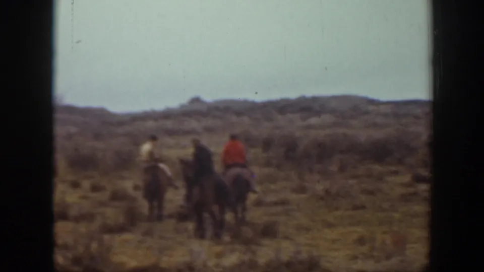 In 1939 Montana home-movie footage, three riders on horseback travel away across sandy open ground with low brush and distant hills.