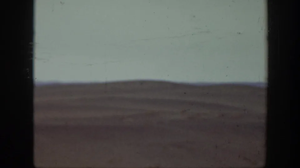 The 8mm frame shows a sparse Montana plain, low horizon, and pale sky, with the riders outside the visible area or too indistinct to identify.