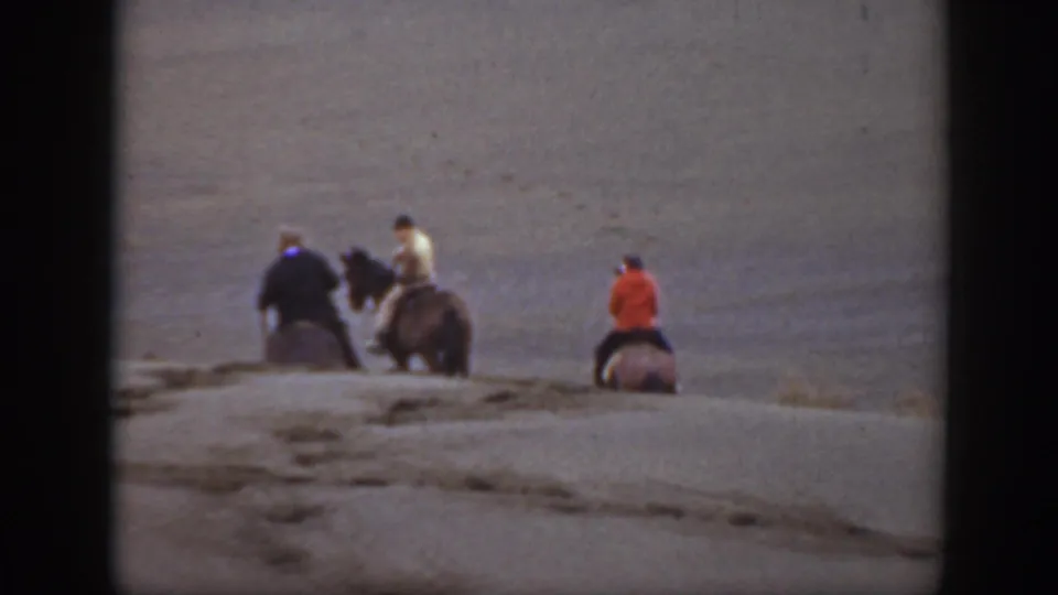 Four distant riders on horseback continue across a bare Montana landscape, with coats and hats visible in soft 8mm color.