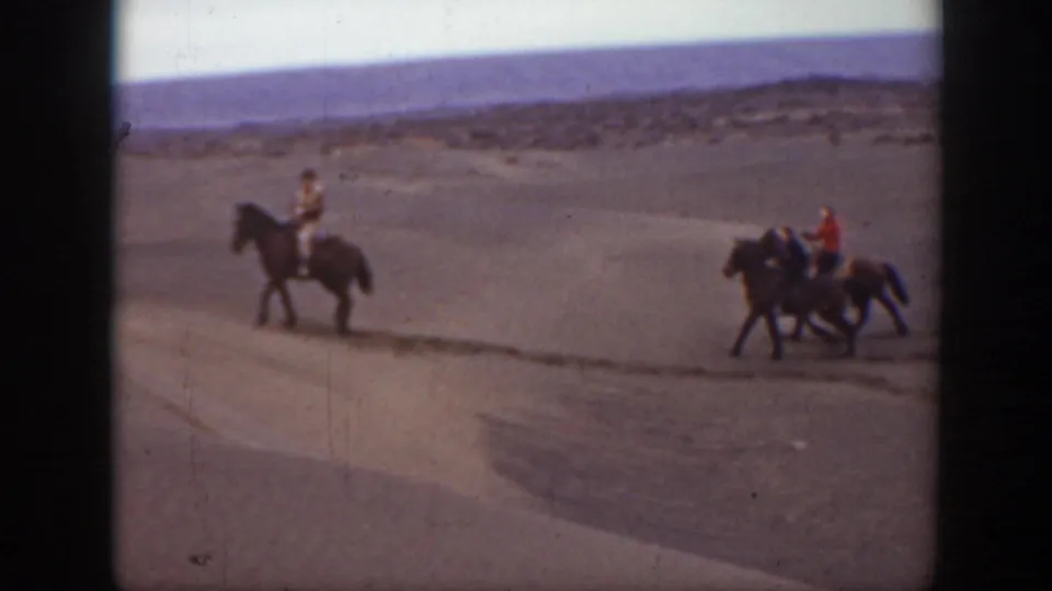 Three riders on horseback appear in the Montana landscape, with one rider ahead and two following across the sandy ground.