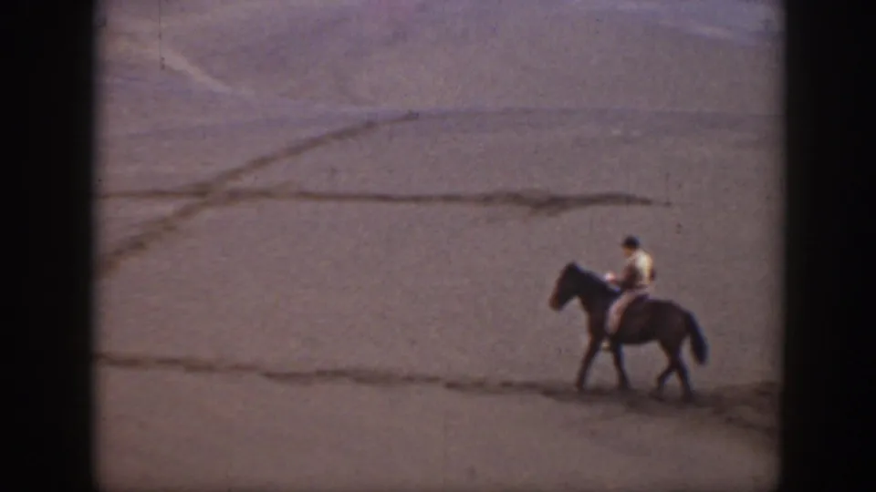 A lone man rides a dark horse across the Montana sand, with faint dune lines and 8mm film softness visible.