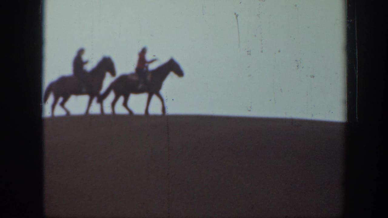 Two horseback riders in silhouette cross a Montana hilltop, framed by pale sky and dark 8mm film edges.
