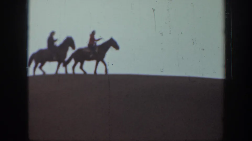 Two horseback riders in silhouette cross a Montana hilltop, framed by pale sky and dark 8mm film edges.
