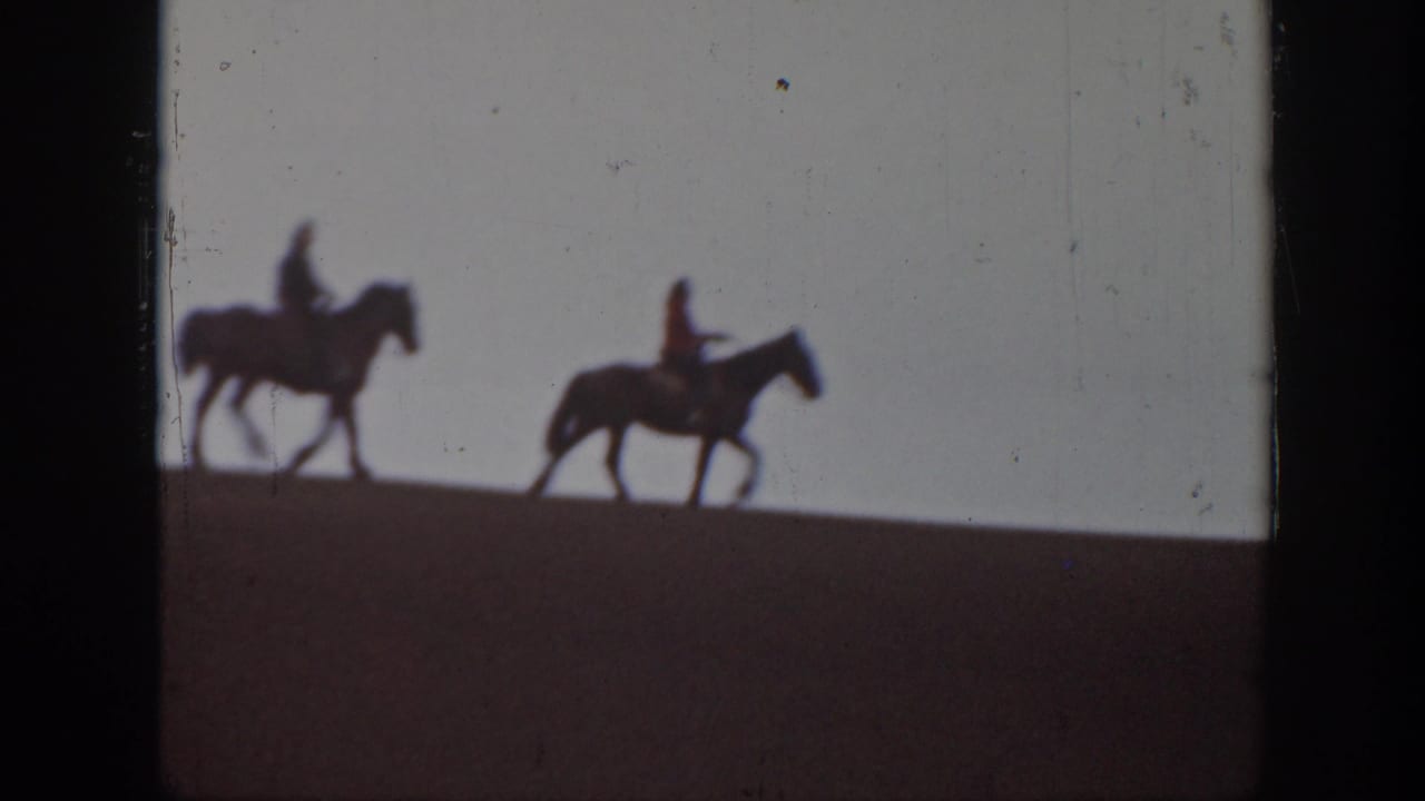 1939 Montana home-movie frame showing two distant horseback riders crossing a sloped sand dune under a pale sky.