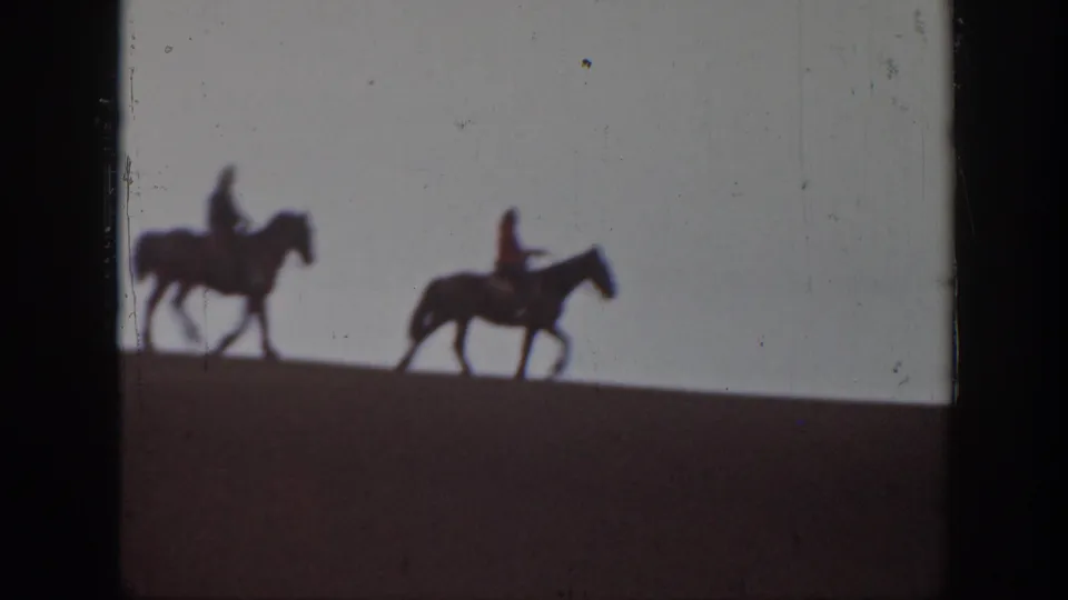 1939 Montana home-movie frame showing two distant horseback riders crossing a sloped sand dune under a pale sky.