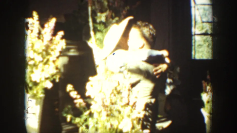 Brief 8mm home-movie frame showing a person indoors beside bright flowers or foliage, with a window and strong overexposure.