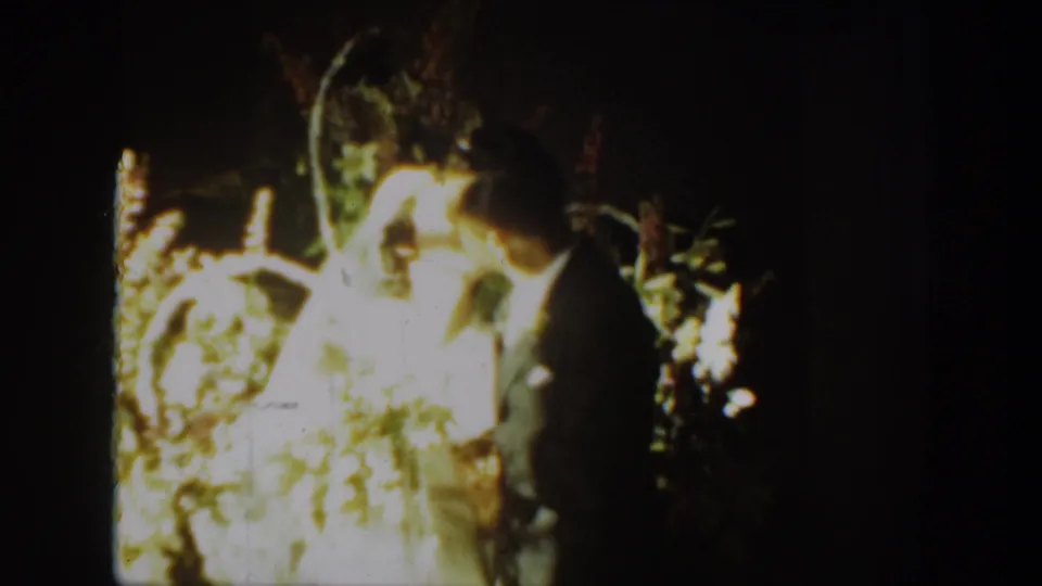 The bride and groom lean into a first-kiss moment in 1939 Montana wedding home-movie footage, with flowers glowing in the background.
