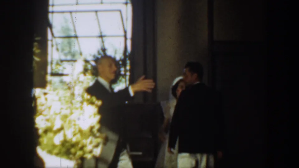 An older man in a suit gestures toward the bride and groom near a window and floral display in the 1939 Montana wedding scene.