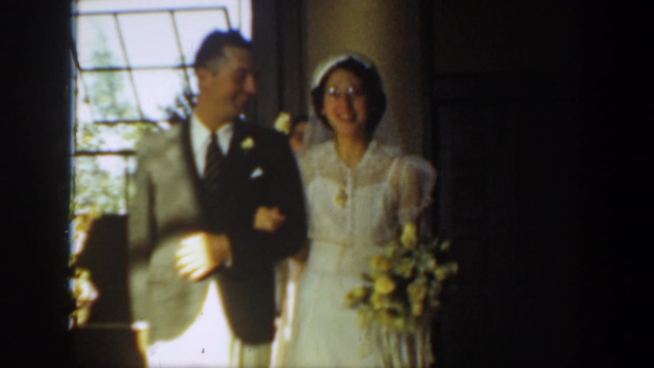 In 1939 Montana home-movie footage, a bride in white walks beside a suited man near a bright window during a wedding procession.