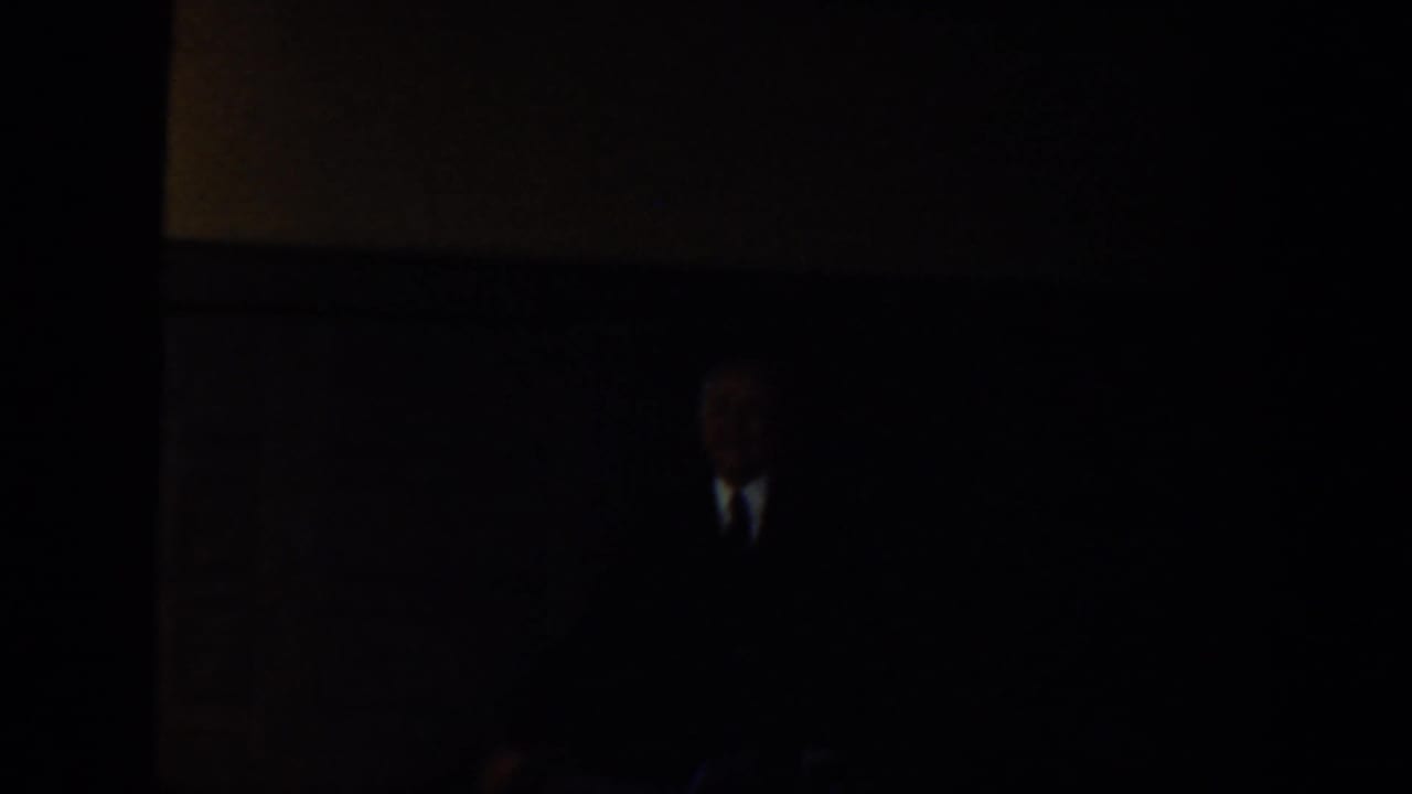 Dim interior with a suited adult figure barely visible near a wall.