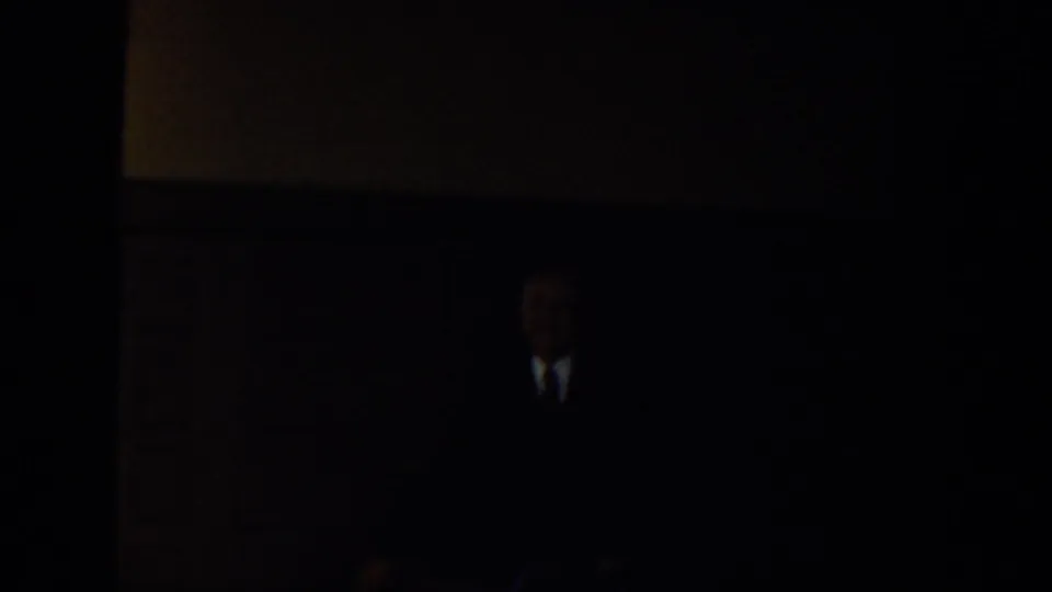 Dim 1939 Montana home-movie interior with a suited adult figure partly visible in low light near a wall or church fixture.
