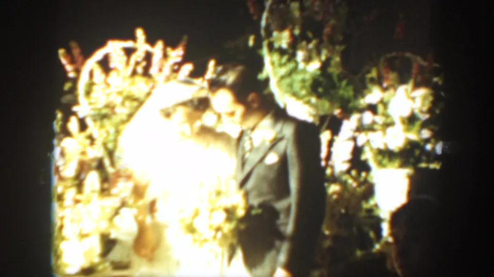Bride in white gown and groom in dark suit stand by an overexposed floral arch in 1939 Montana wedding home-movie footage.