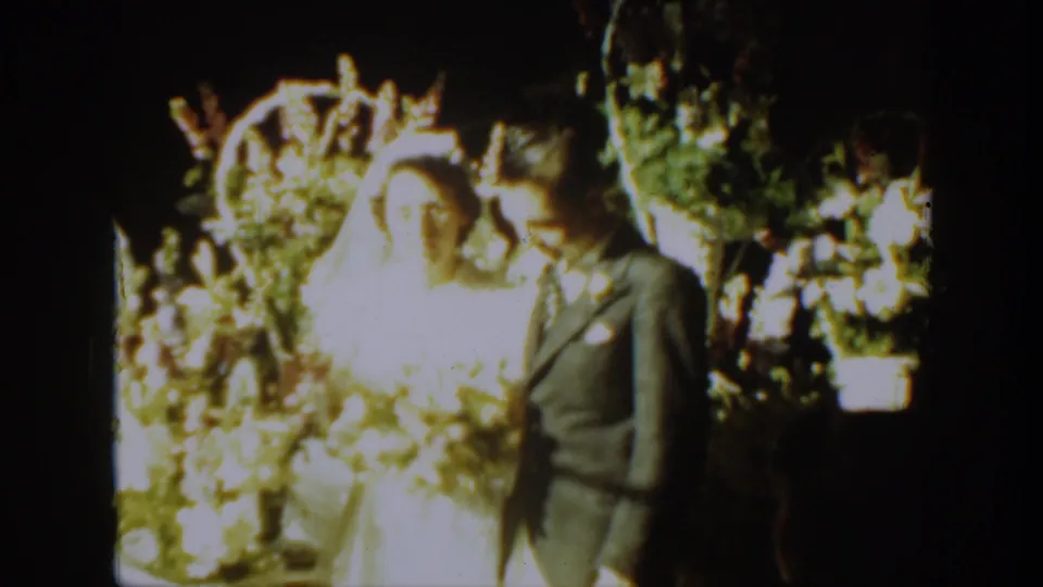 1939 Montana wedding home-movie frame of a bride in veil and groom in suit posing before a floral arch and bouquets.