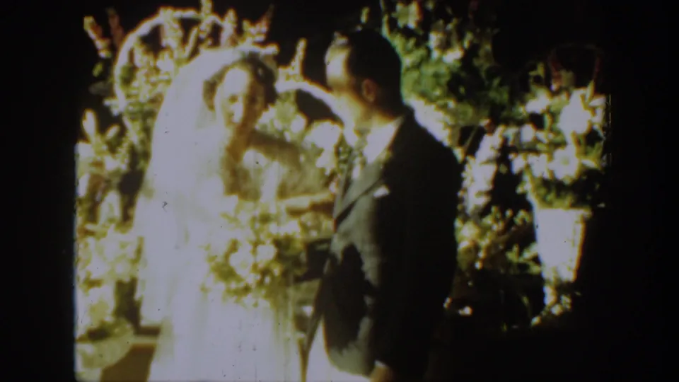 The groom looks toward the bride as she holds a bouquet, framed by wedding flowers in the 1939 Montana home movie.