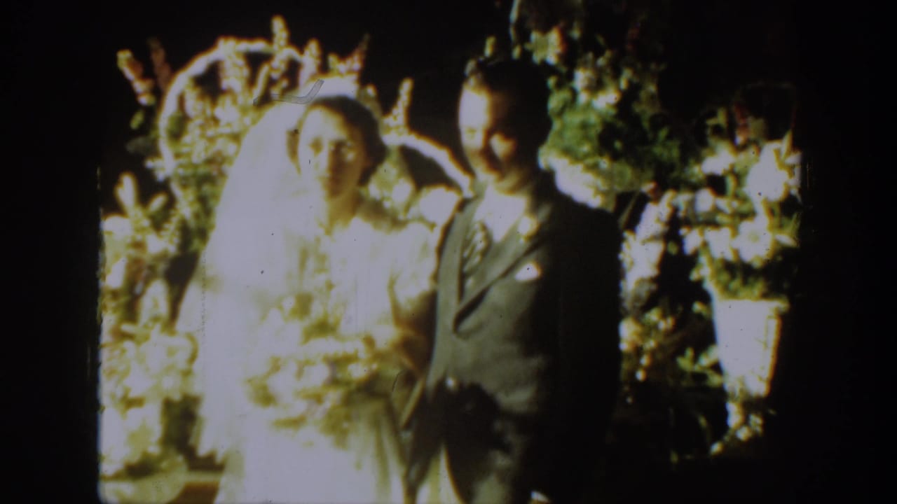 1939 Montana 8mm frame of a bride in a white veil beside a groom in a dark suit before flowers.