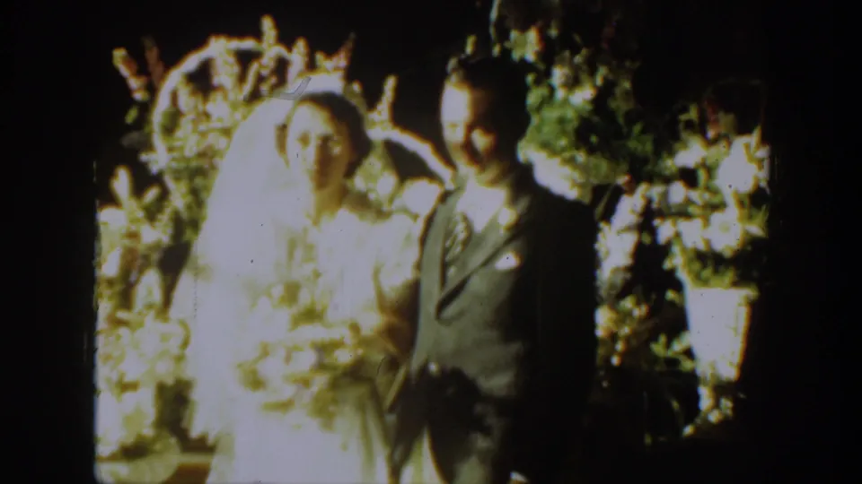 1939 Montana 8mm frame of a bride in a white veil beside a groom in a dark suit before flowers.