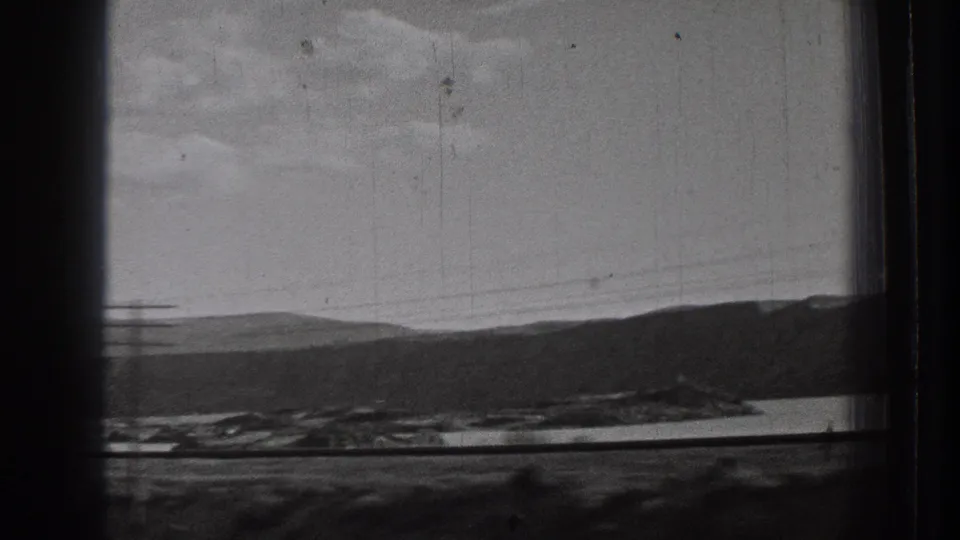 1939 Washington 8mm frame showing a rail or road corridor along water, with low hills, poles, and cloudy sky in the distance.