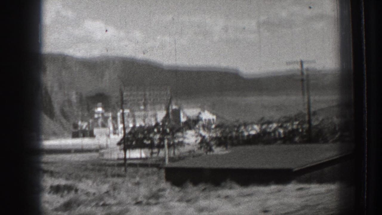 Roadside view of a dam sign and industrial structures with hills behind.