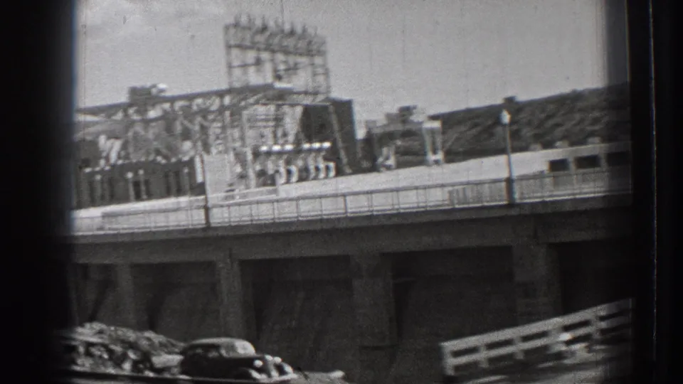 1939 Washington frame shows dam-area industrial structures, an elevated bridge or road, railings, a dark car, and hills beyond.