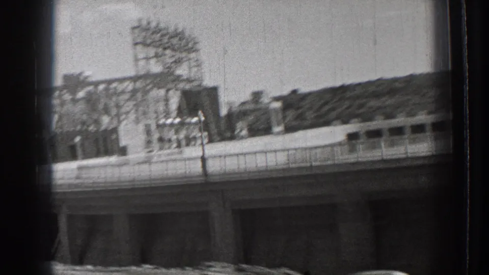 1939 Washington 8mm frame showing a drive-by view of a dam approach with railing, industrial framework, water, and a hillside.