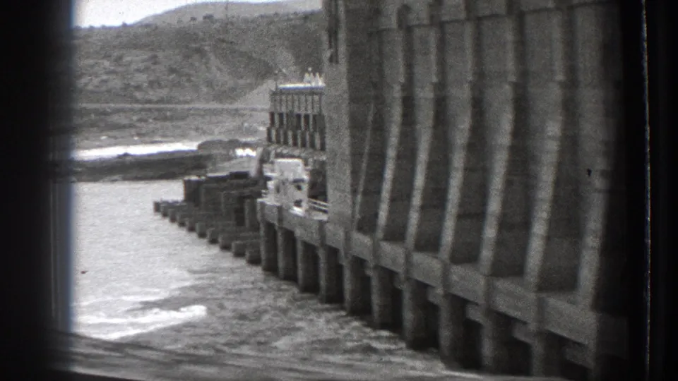 1939 Washington 8mm frame looking along a large concrete dam wall toward gate structures, flowing water, and hills.