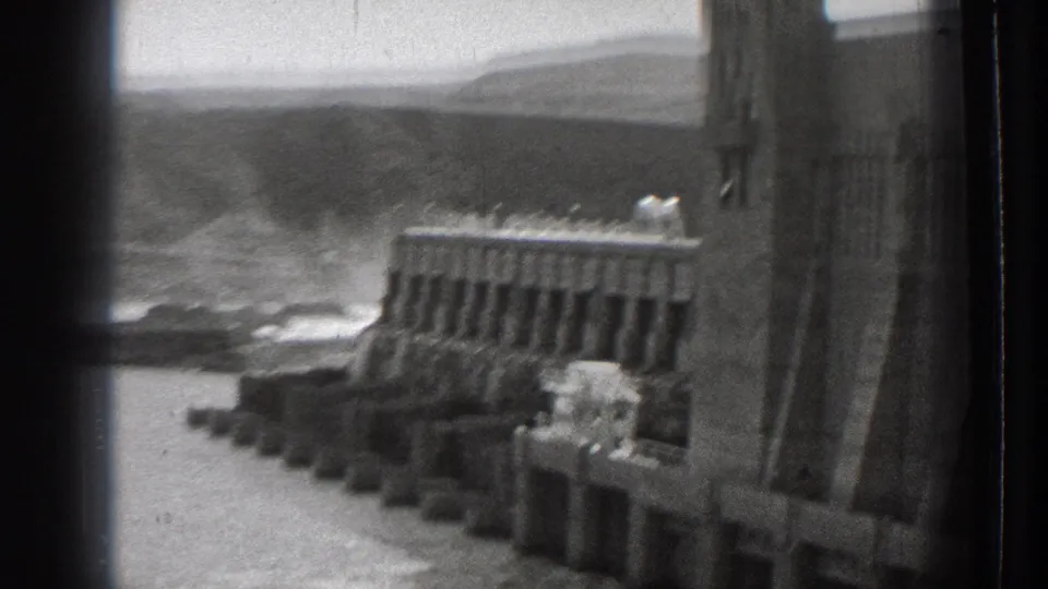 1939 Washington 8mm frame showing the dam wall at right, spillway structures, water below, and hazy hills in the background.