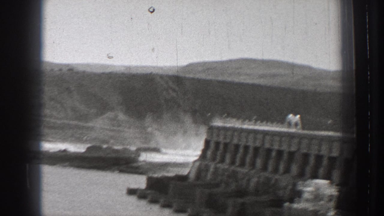 Waves crash near a concrete breakwater with low hills in the background.