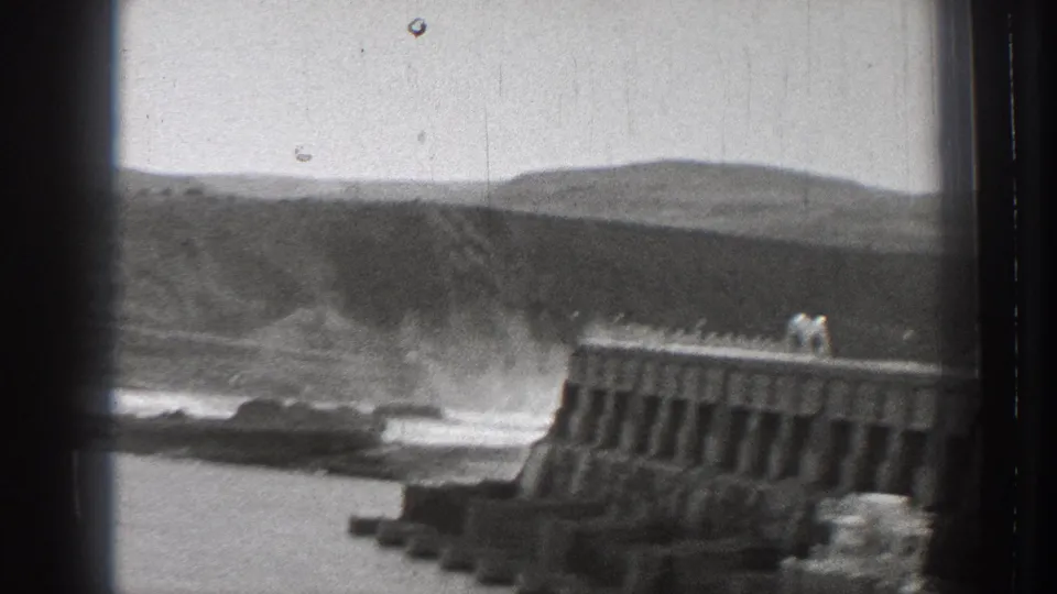 1939 Washington 8mm frame showing surf breaking beside a concrete breakwater, with low hills beyond the shoreline.