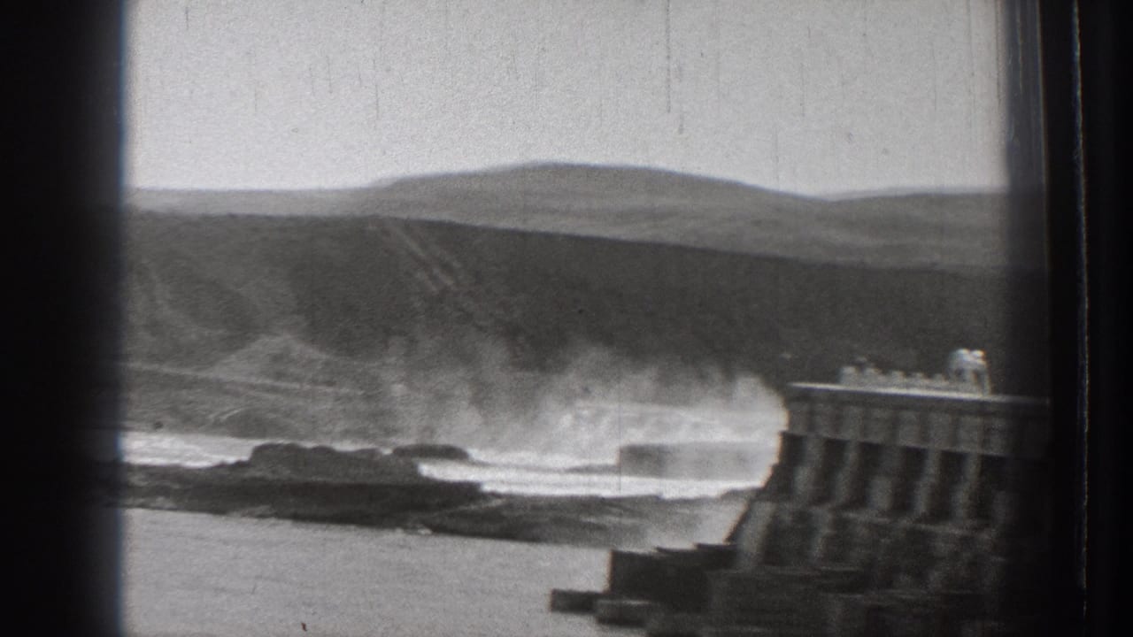 Spray rises beside a concrete breakwater with hills behind the shore.