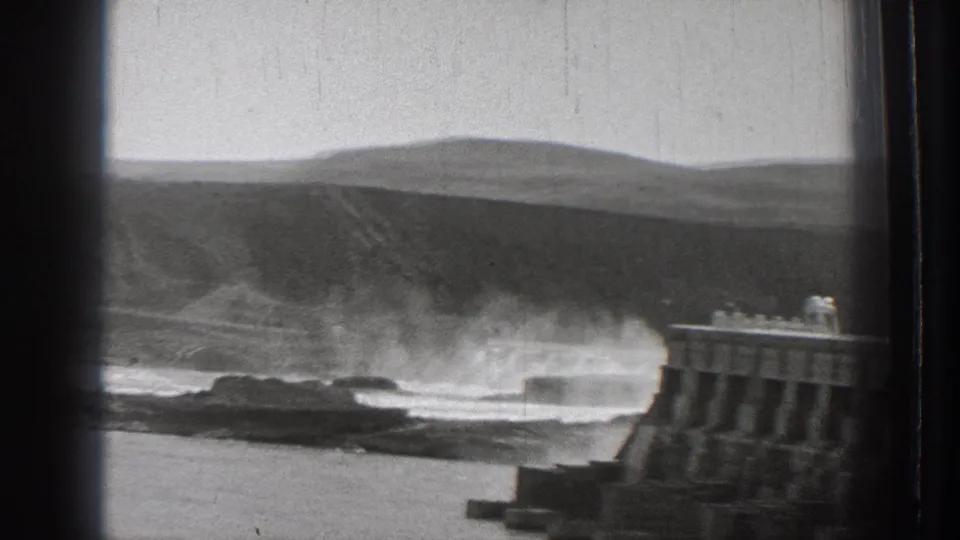 1939 Washington coastal frame with wave spray hitting the breakwater and open hills behind the waterline.