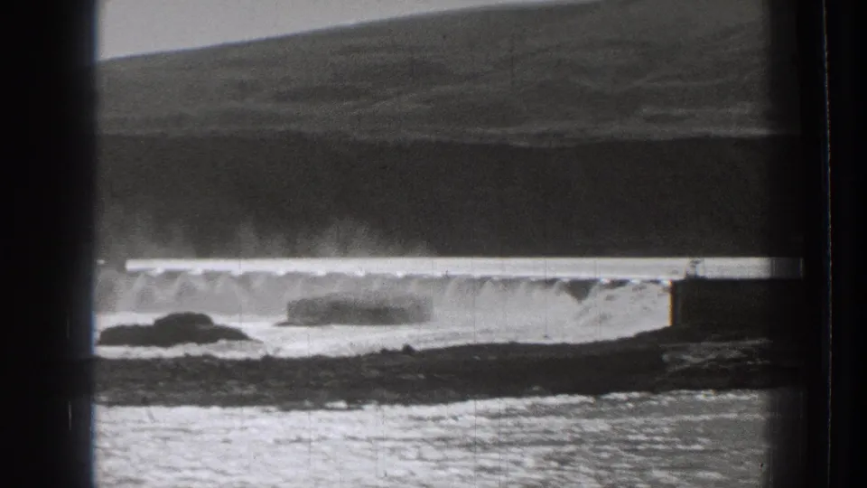1939 Washington 8mm frame showing white surf along a low coastal structure, with grassy hills and rocks visible.