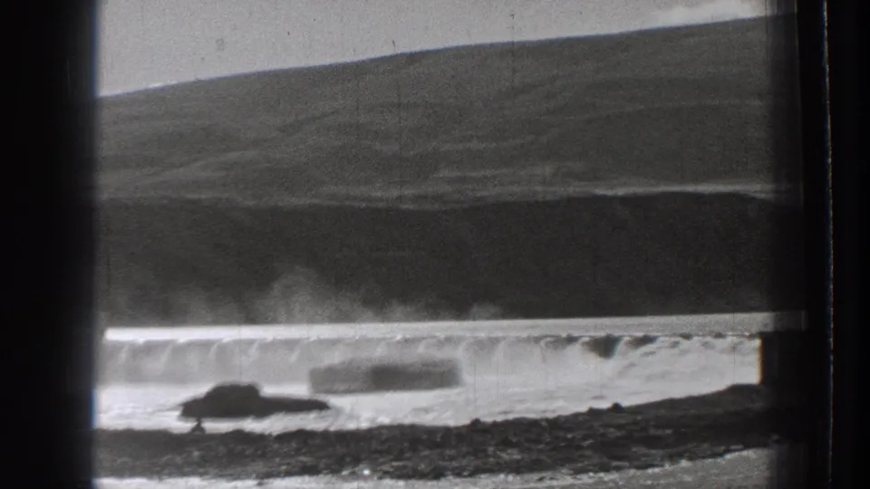 1939 Washington coastal home-movie frame showing surf breaking along a low wall and rocky foreground under open hills.