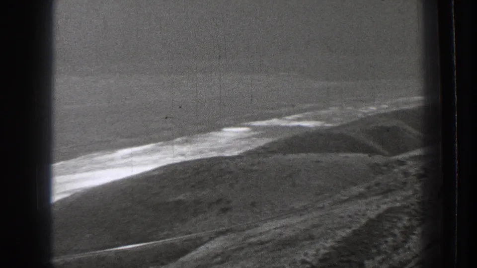 Black-and-white 8mm frame from Washington showing low waterfront terrain and pale surf from a train-window perspective.