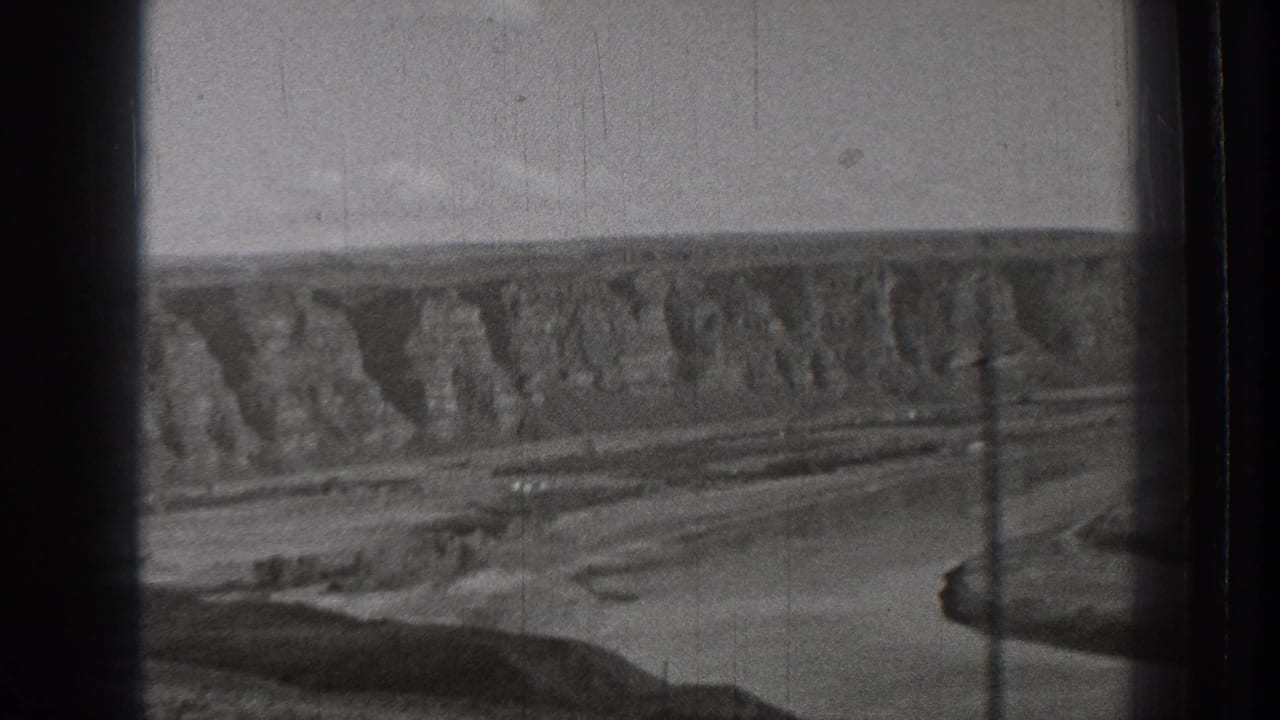 Rugged cliffs, open ground, utility poles, and a winding channel in black-and-white footage.