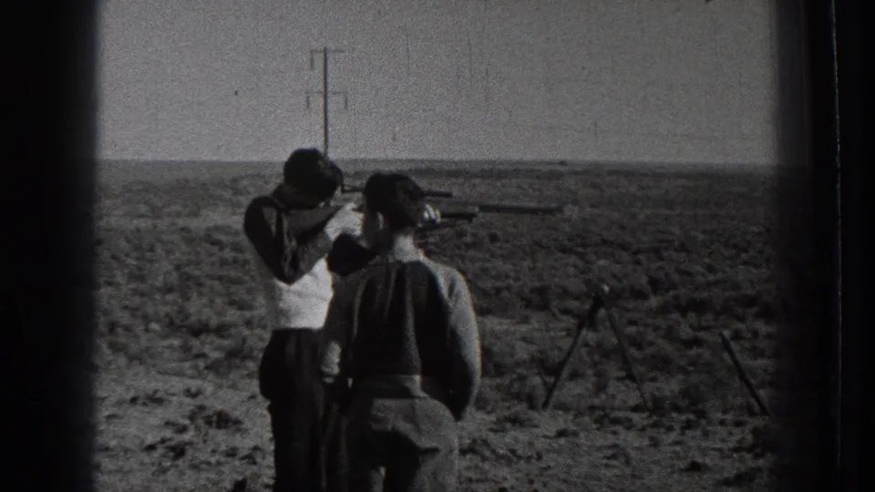 Two men in 1939 Washington wilderness footage stand in scrubland, one holding a rifle near his shoulder as utility poles cross the background.