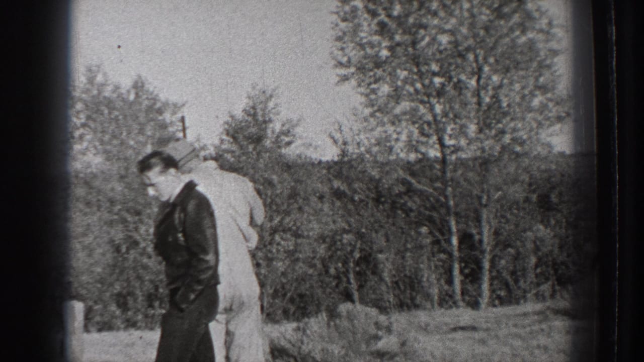 Two adult men stand in a field near trees, one in a dark jacket and one in a light suit.