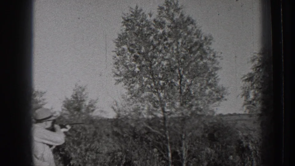 1939 Washington home-movie frame of an adult man aiming a firearm in a rural field beside a large tree and distant horizon.