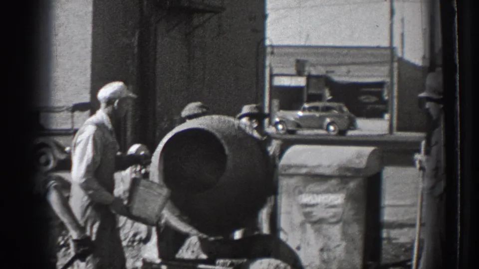 A worker in cap and work clothes handles a bucket beside a cement mixer, with another worker and a parked car visible near the street.