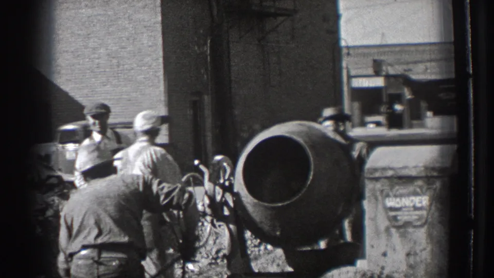 At the construction site, one worker bends toward materials while others stand near the cement mixer, with a marked bin and street behind them.