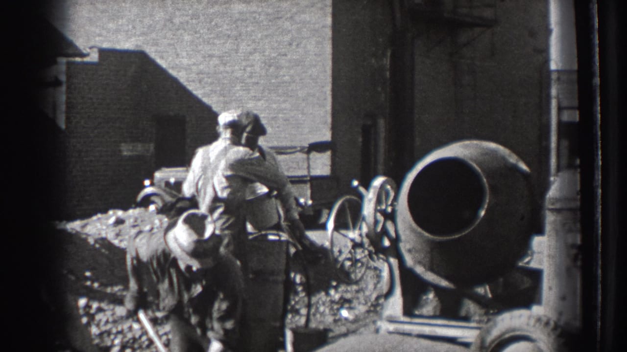 Workers shovel material near a cement mixer with a vintage automobile in the background.