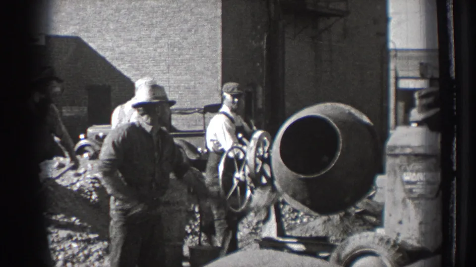 A worker in a brimmed hat crosses in front of the cement mixer as other men stand near the machinery and material pile.