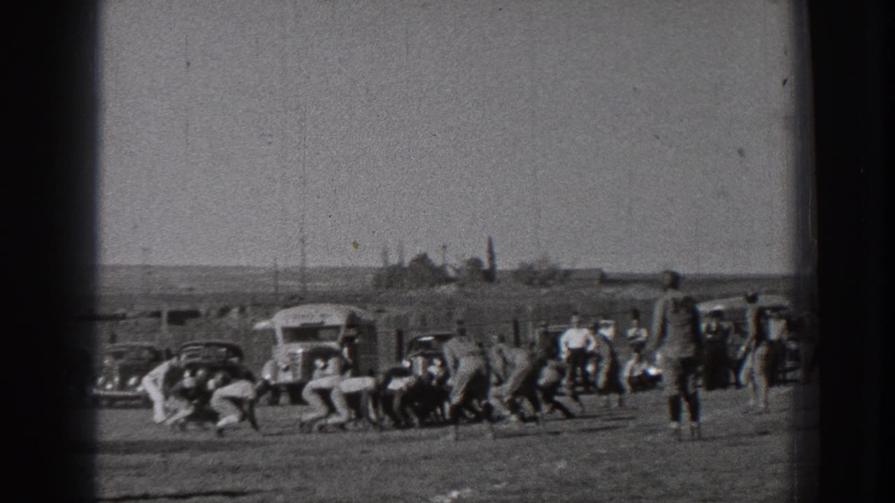 1939 Washington home-movie frame showing football players near the line of play on a dusty field with cars, a bus, and spectators in the background.