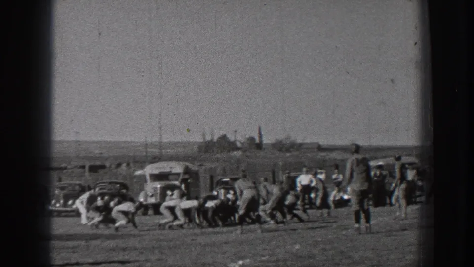 1939 Washington home-movie frame showing football players near the line of play on a dusty field with cars, a bus, and spectators in the background.