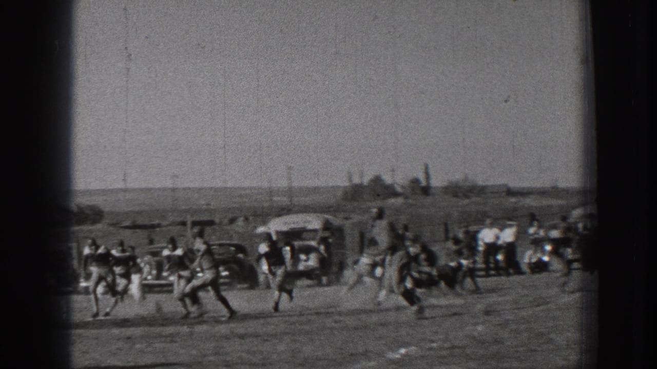 Football players run across a dirt field as spectators and vehicles line the background.
