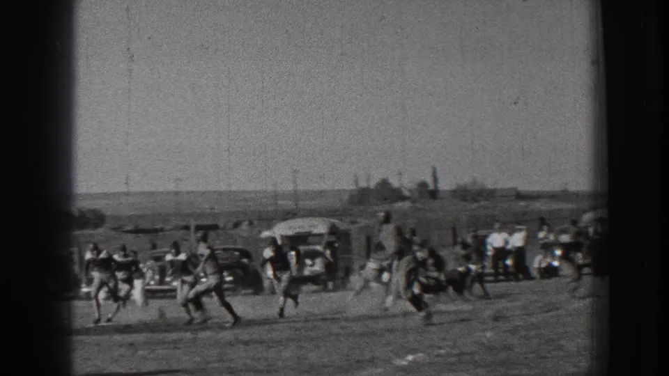 Football players in 1939 Washington run during a play on an open dirt field, with parked cars, a bus, and sideline onlookers behind them.