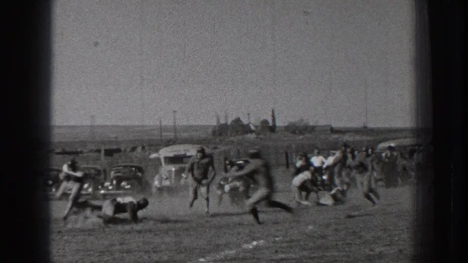 A player appears down on the dirt during a 1939 Washington football play, with other players moving through dust and spectators near vehicles behind them.