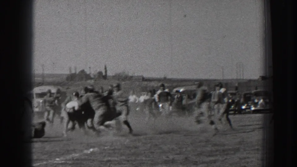 Football players converge in a dusty tackle on a Washington dirt field in 1939, with spectators and old-style cars visible along the sideline.