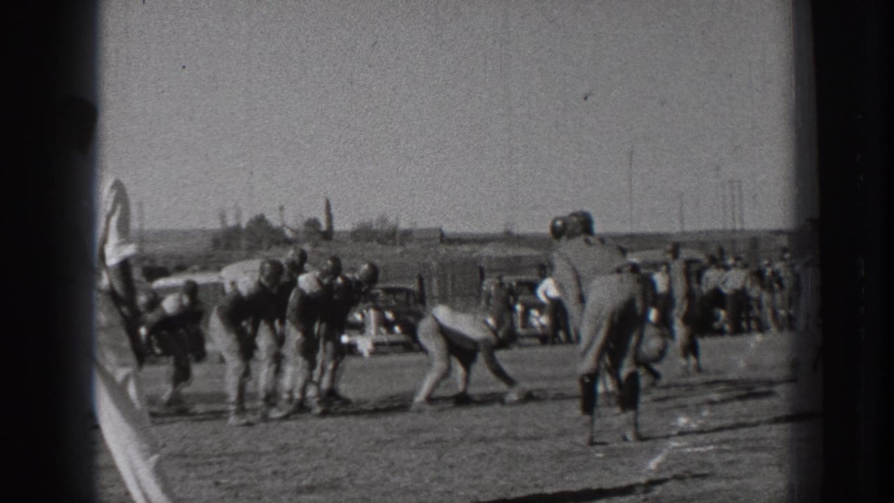 1939 Washington home-movie frame showing young football players preparing at the scrimmage line on a grassy field, with spectators and parked period cars in the background.