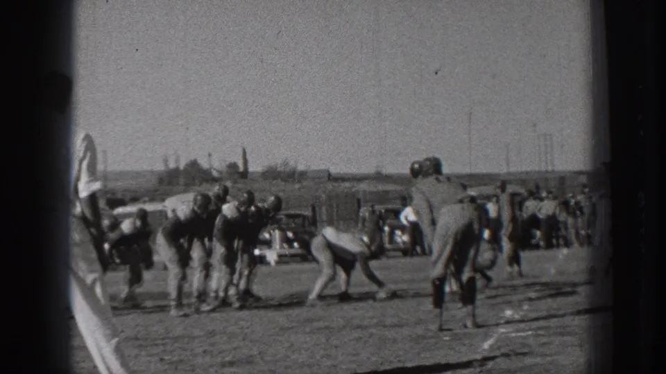 1939 Washington home-movie frame showing young football players preparing at the scrimmage line on a grassy field, with spectators and parked period cars in the background.