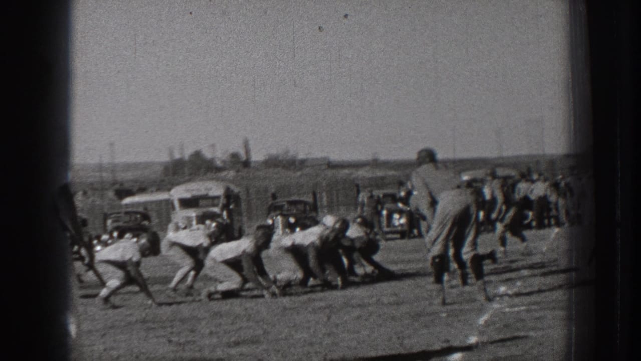Football players crouch and begin a play while cars and spectators stand along the far side of the field.